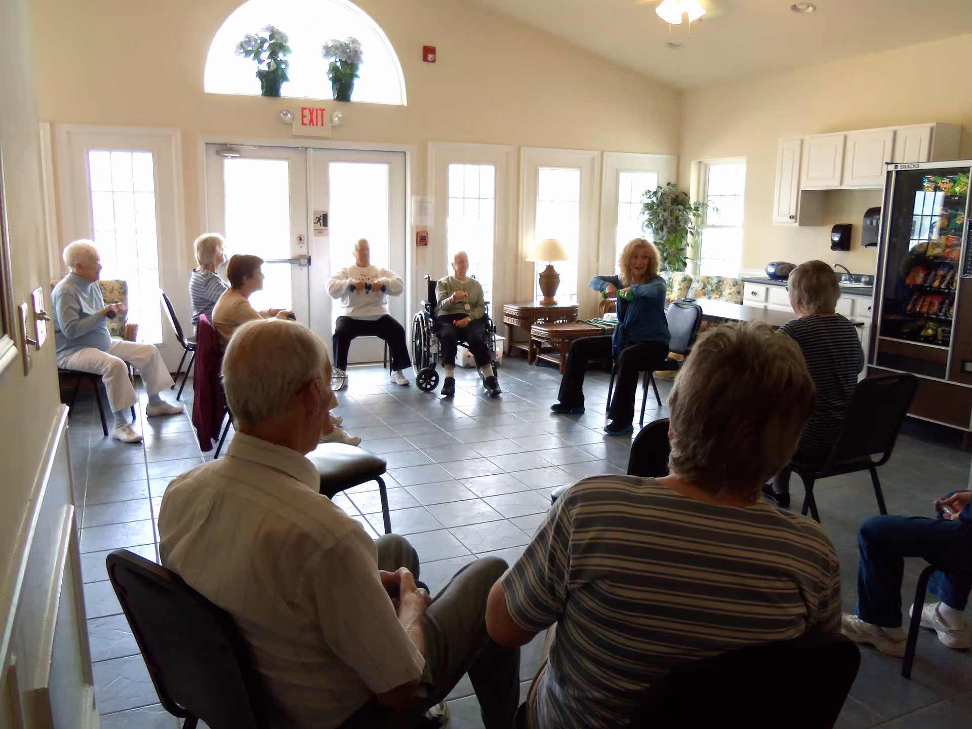 A group of elderly people seated in a circle in a bright room with large windows and a glass door. Some participants are holding small weights and appear to be engaged in a seated exercise or activity. The room has tiled flooring, a vending machine, a small kitchenette area, and a lamp on a side table.