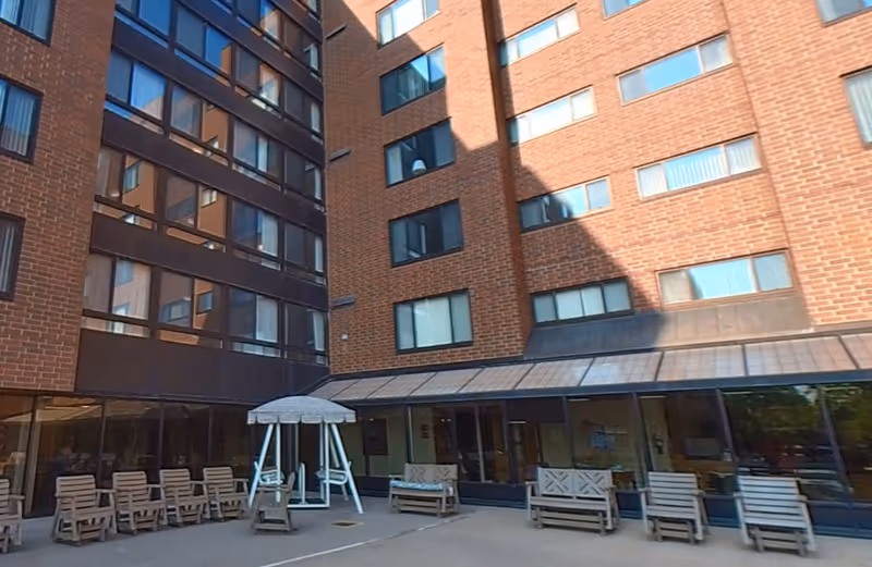 Outdoor courtyard area of a senior living facility with multiple wooden chairs and benches arranged around a concrete patio. The building surrounding the courtyard is made of red brick with many windows. There is a white canopy swing in the center of the patio.