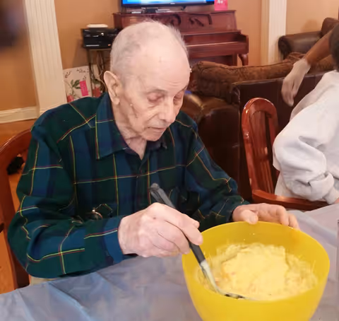 An elderly man wearing a green plaid shirt is sitting at a table and stirring a yellow bowl filled with a creamy mixture. In the background, there is a piano, a television, and a brown couch in a cozy living room setting.