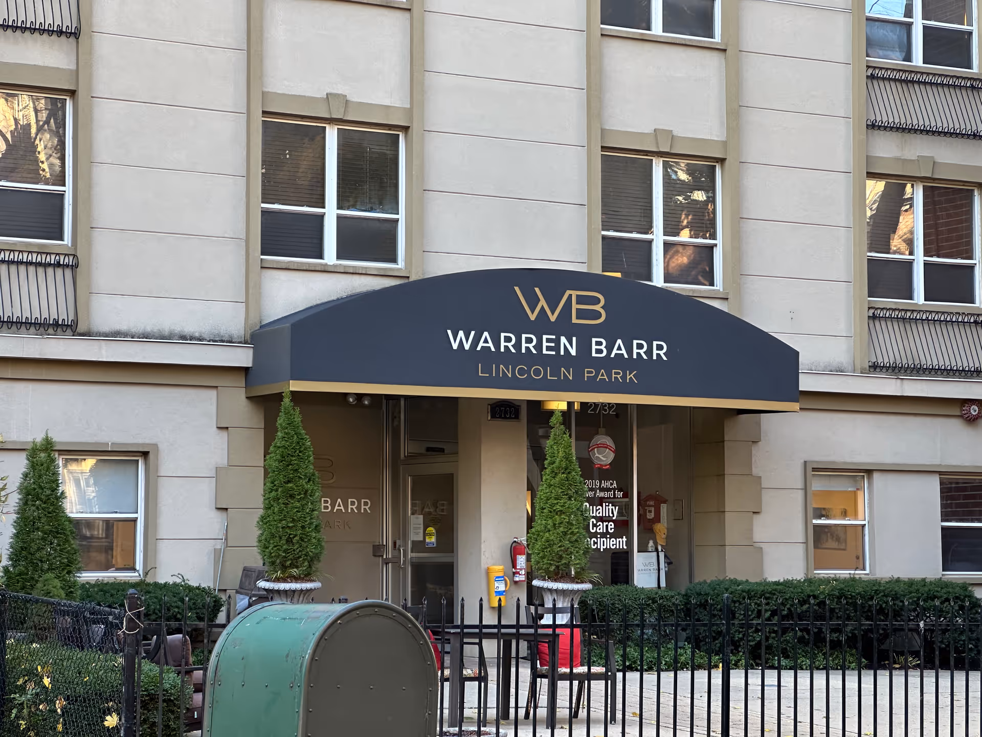 Front entrance of the Warren Barr Lincoln Park building with a black awning bearing the WB logo, flanked by potted evergreens and a black fence.