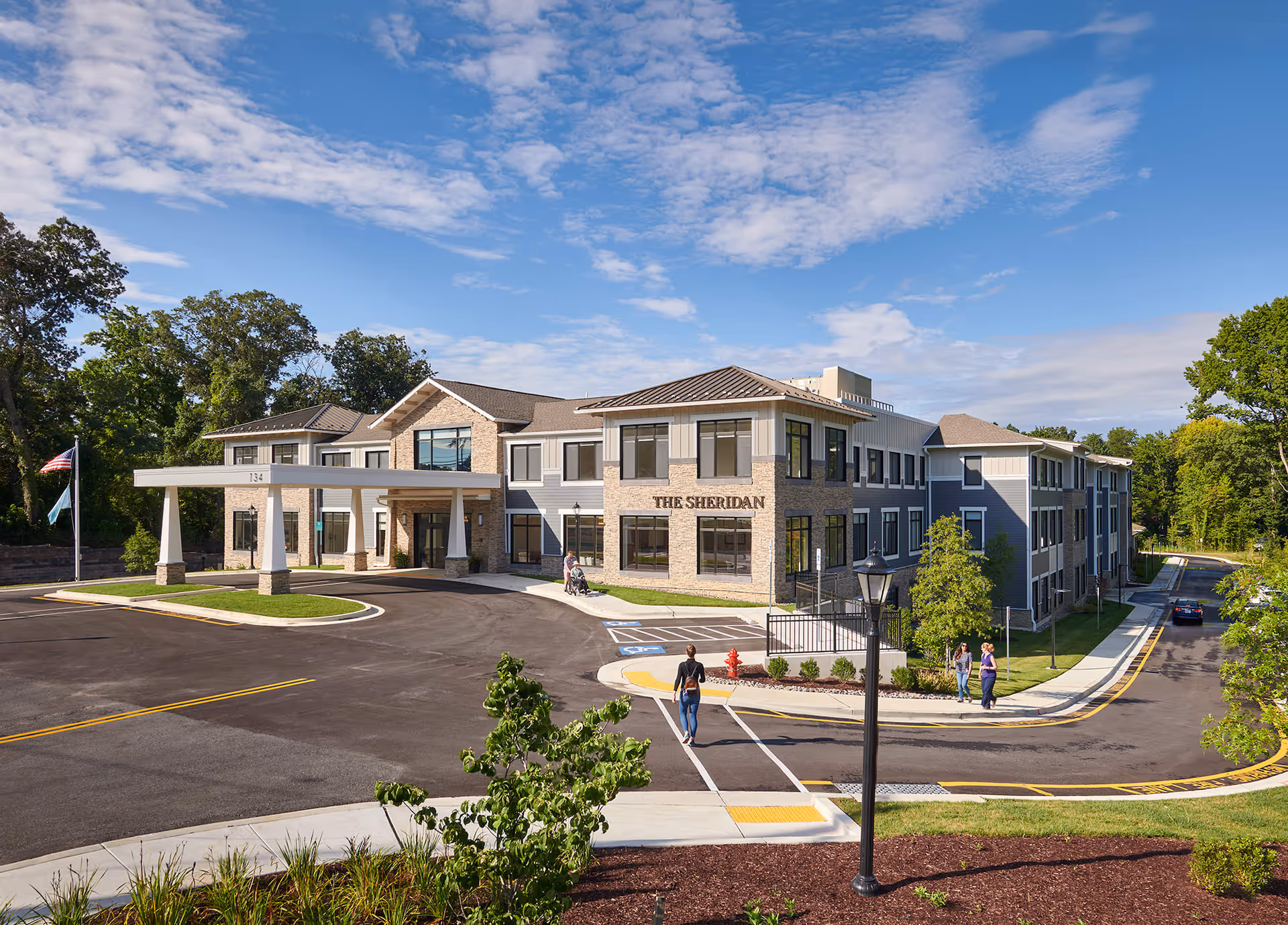 Exterior view of The Sheridan At Severna Park, a modern two-story senior living facility with a covered entrance, parking spaces, and landscaped surroundings under a partly cloudy blue sky. Several people are walking near the entrance and along the sidewalk.