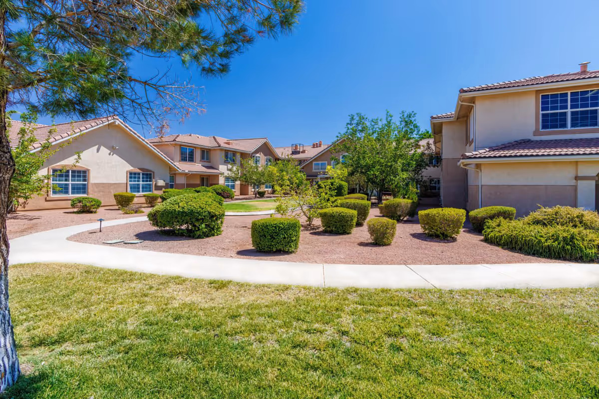 Sunny landscaped courtyard with trimmed shrubs, walkways, and beige two-story assisted living buildings under a clear blue sky.
