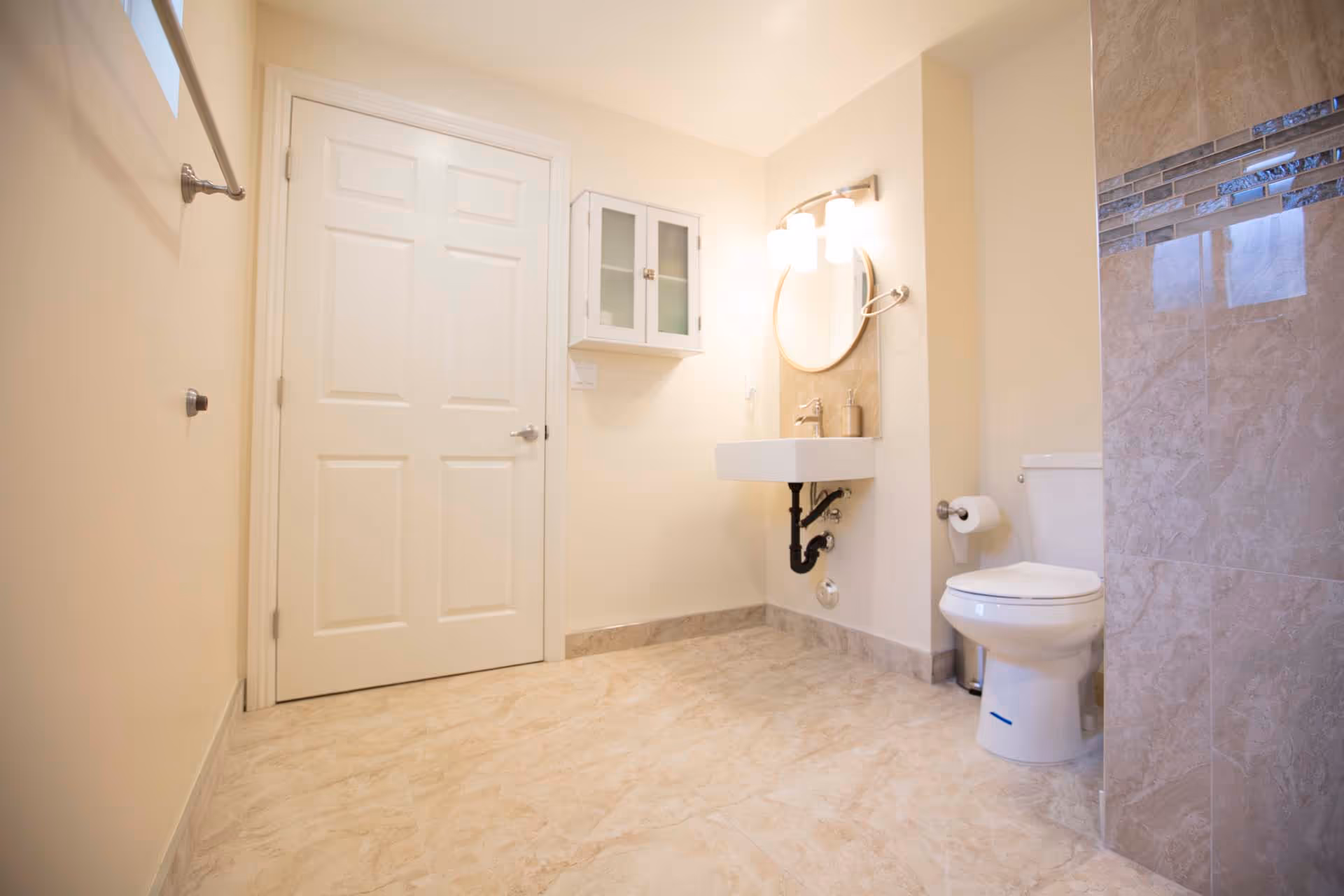 Bright, modern bathroom with a wall-mounted sink and round mirror, a toilet, and tiled floors.