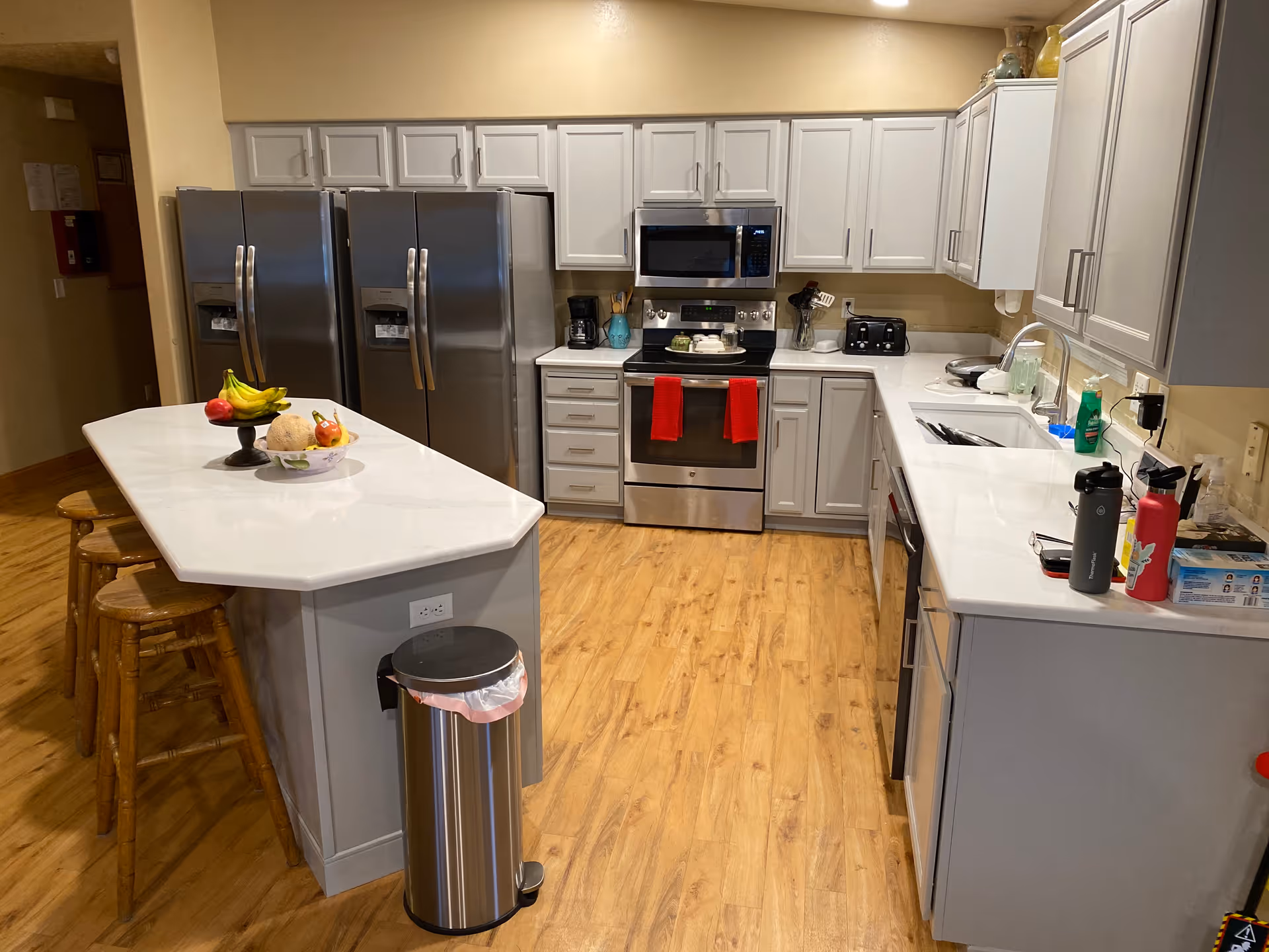 A modern kitchen with light gray cabinets, stainless steel appliances including two refrigerators, an oven with a microwave above it, and a dishwasher. The kitchen has a white countertop island with three wooden stools, a fruit bowl, and a trash can beside it. The floor is wooden, and various kitchen items like a coffee maker, toaster, and water bottles are visible on the counters.