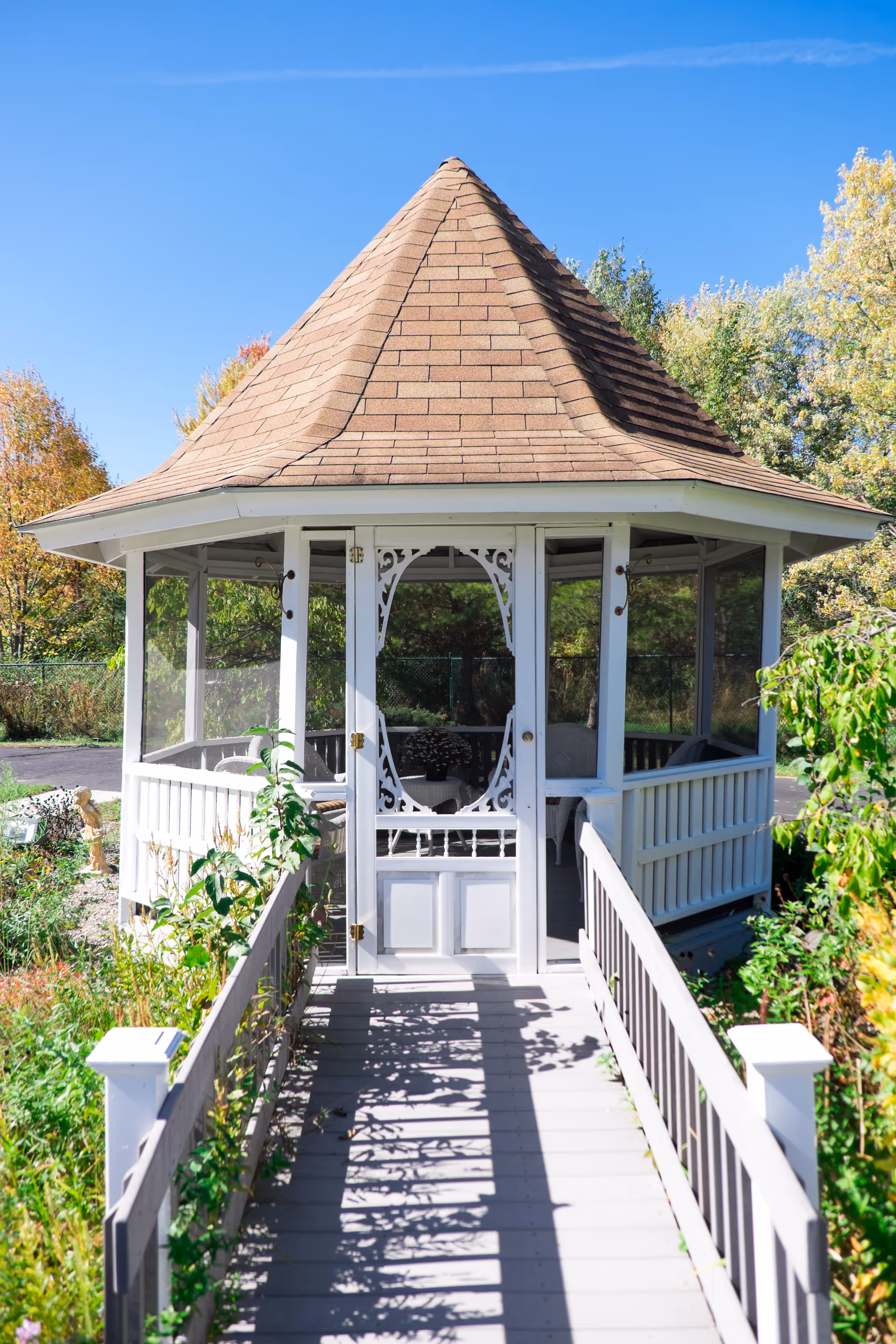 A white wooden gazebo with a shingled roof and screened windows, situated at the end of a wooden walkway surrounded by greenery and plants under a clear blue sky.