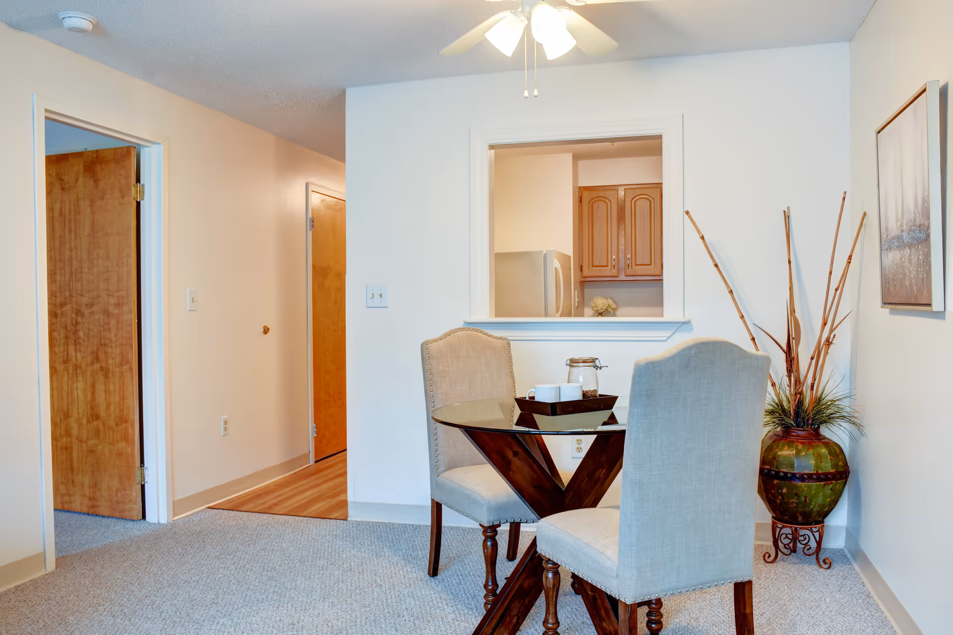 A small dining area with a round glass-top table and two upholstered chairs. The table has a tray with a jar and two cups. Behind the table is a pass-through window to a kitchen with wooden cabinets and a refrigerator. To the right of the table is a large decorative vase with tall dried branches and a framed artwork on the wall. The floor is carpeted and there is a ceiling fan with lights above.