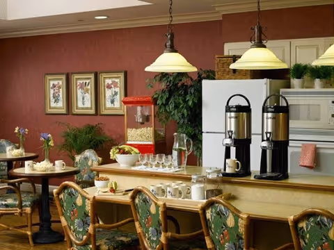 Interior view of a dining area with a counter featuring coffee dispensers, cups, and a popcorn machine. There are floral upholstered chairs around tables and the counter. The walls are decorated with framed floral artwork and there are hanging pendant lights above the counter.