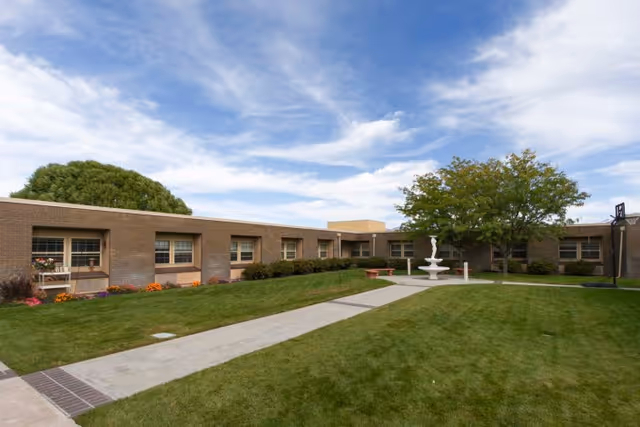 Well-maintained grassy courtyard with a walkway leading to a central fountain, surrounded by a single-story brick health care building and trees.