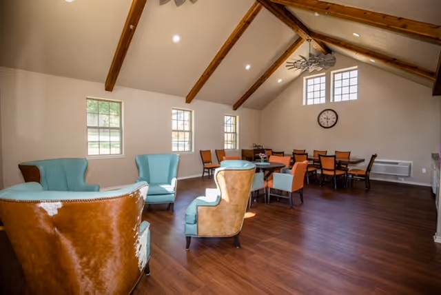 Sunlit communal lounge and dining area with teal and brown armchairs, dining tables, exposed wooden beams, and a wall clock.