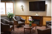A cozy living room area with four brown leather armchairs arranged around a wooden coffee table with a vase of flowers. There is a large flat-screen TV mounted on the wall above a wooden fireplace. A window with curtains and a green plant is visible on the left side.