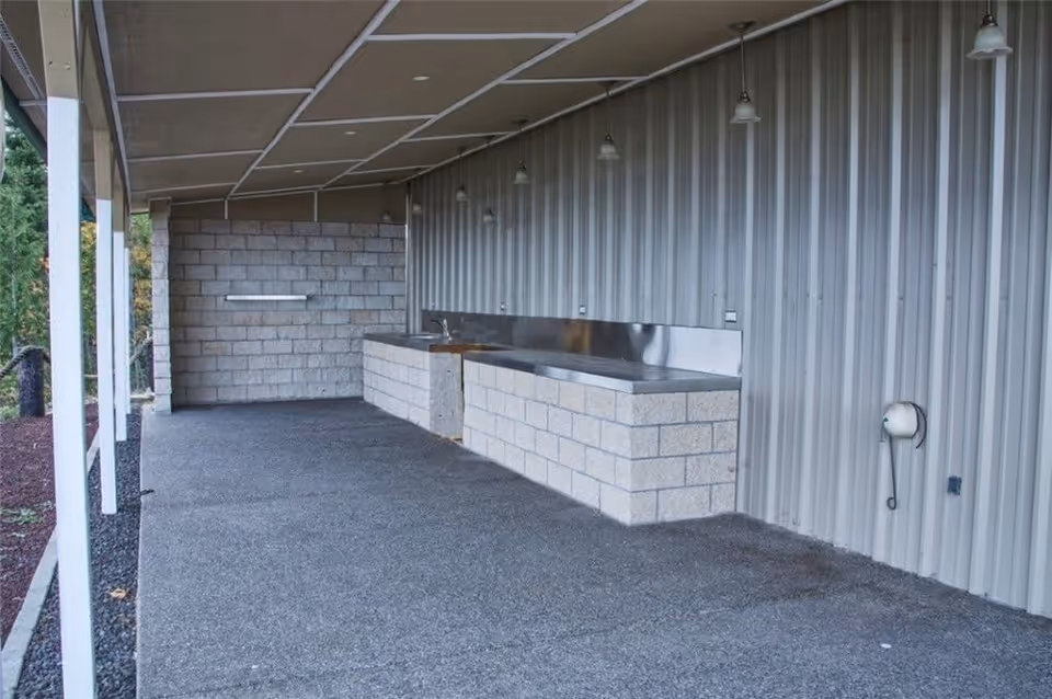 Covered outdoor area with a concrete floor and a long countertop made of concrete blocks and stainless steel. The wall behind the countertop is made of corrugated metal, and there are several hanging lights above the countertop. The area is supported by white pillars and overlooks some greenery.