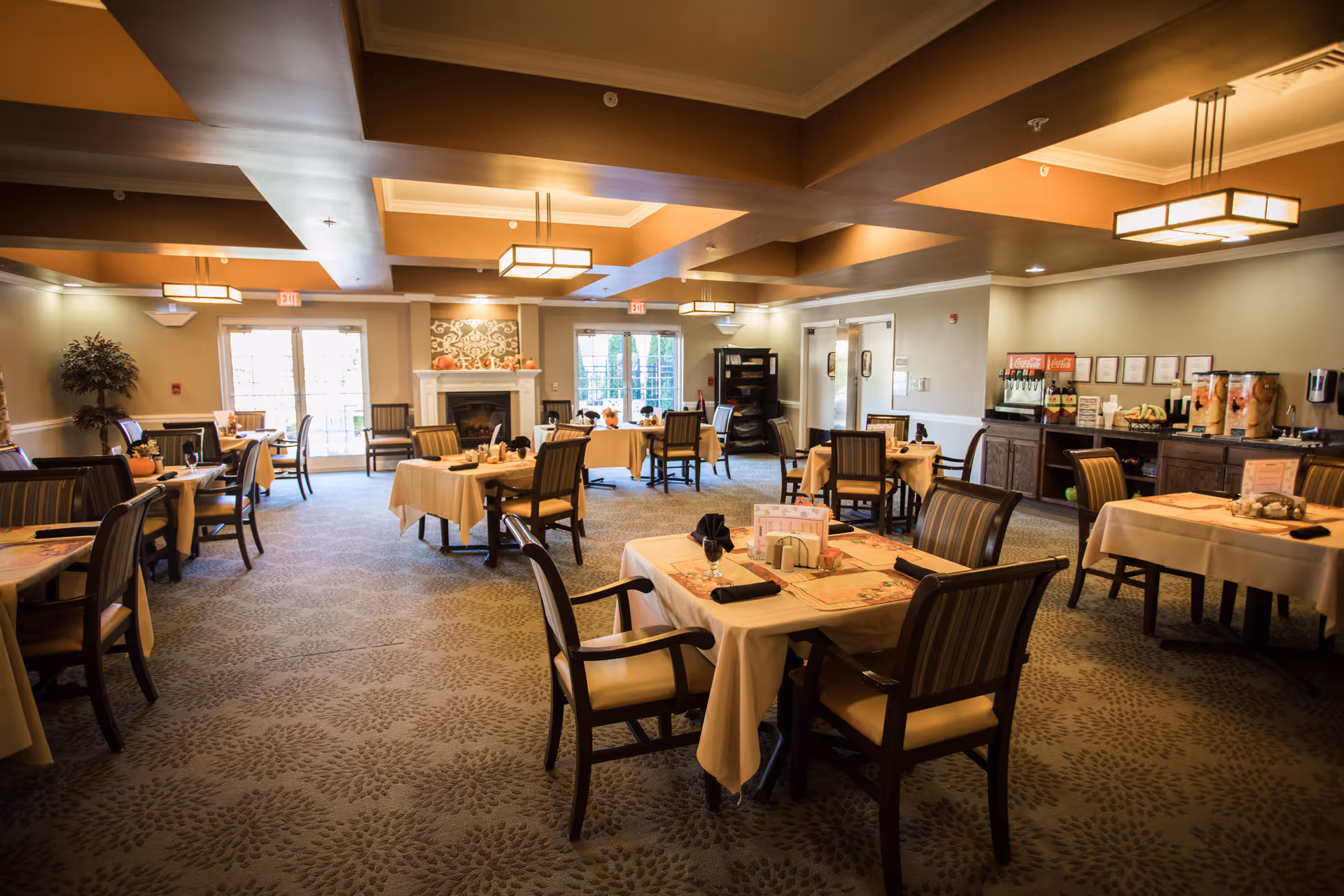 A spacious dining room with multiple tables covered in beige tablecloths, each set with napkins and menus. The room features patterned carpet, a fireplace with decorative artwork above it, and large windows letting in natural light. There is a beverage station with soda dispensers and fruit on the right side of the room.