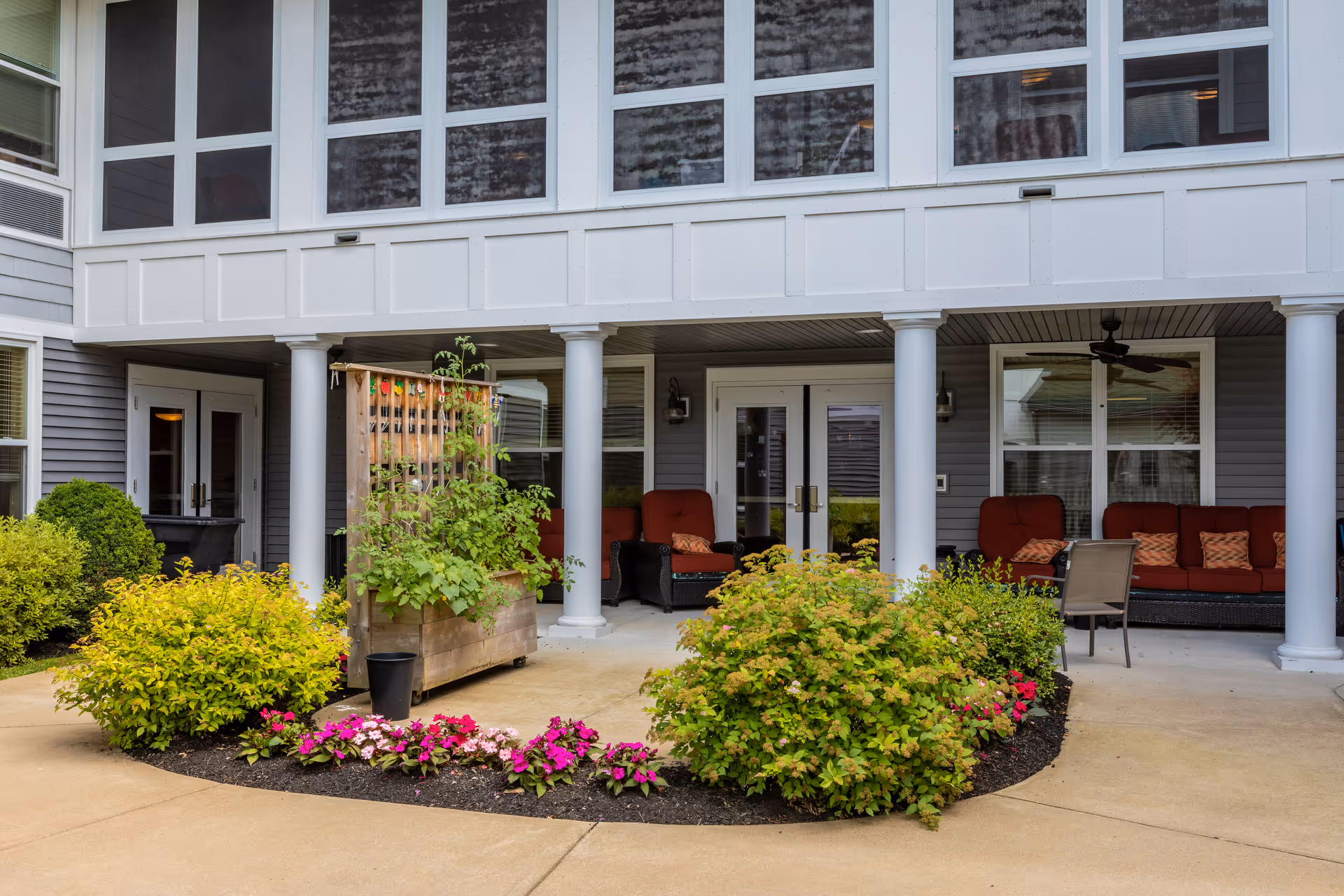 Outdoor patio area at The Inn at Fairview Memory Care featuring a covered seating space with red cushioned chairs and a sofa, white columns, and a garden bed with green shrubs and colorful flowers in front.