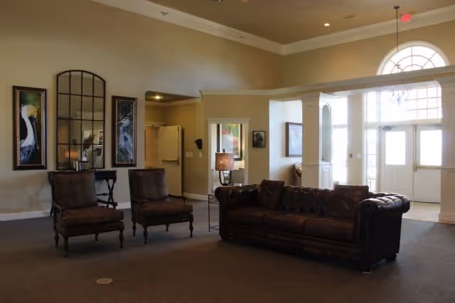 A spacious living room area with a dark brown leather sofa and two matching armchairs. The room features beige walls with two framed bird paintings and a large decorative mirror. There is a lamp on a side table next to the sofa, and large windows and a glass door letting in natural light.