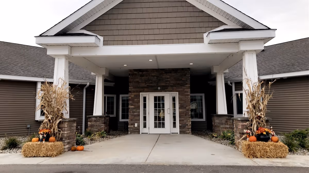 Entrance of Oscoda Fields Assisted Living & Memory Care facility with a covered porch supported by white columns. The entrance features double glass doors framed by stone pillars. Seasonal decorations including hay bales, pumpkins, and dried corn stalks are placed on either side of the walkway.