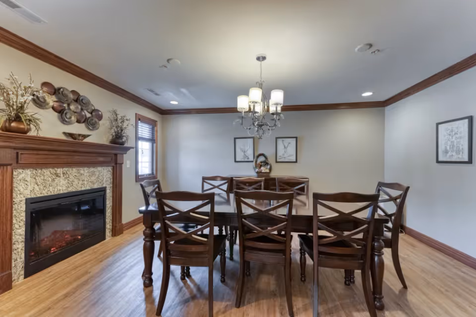 A dining room with a wooden dining table and six matching chairs. There is a decorative fireplace on the left wall with a mantel adorned with plants and wall art above it. The room has wooden flooring, crown molding, and a chandelier hanging from the ceiling. Two framed pictures hang on the back wall, and a window with blinds is on the left side.