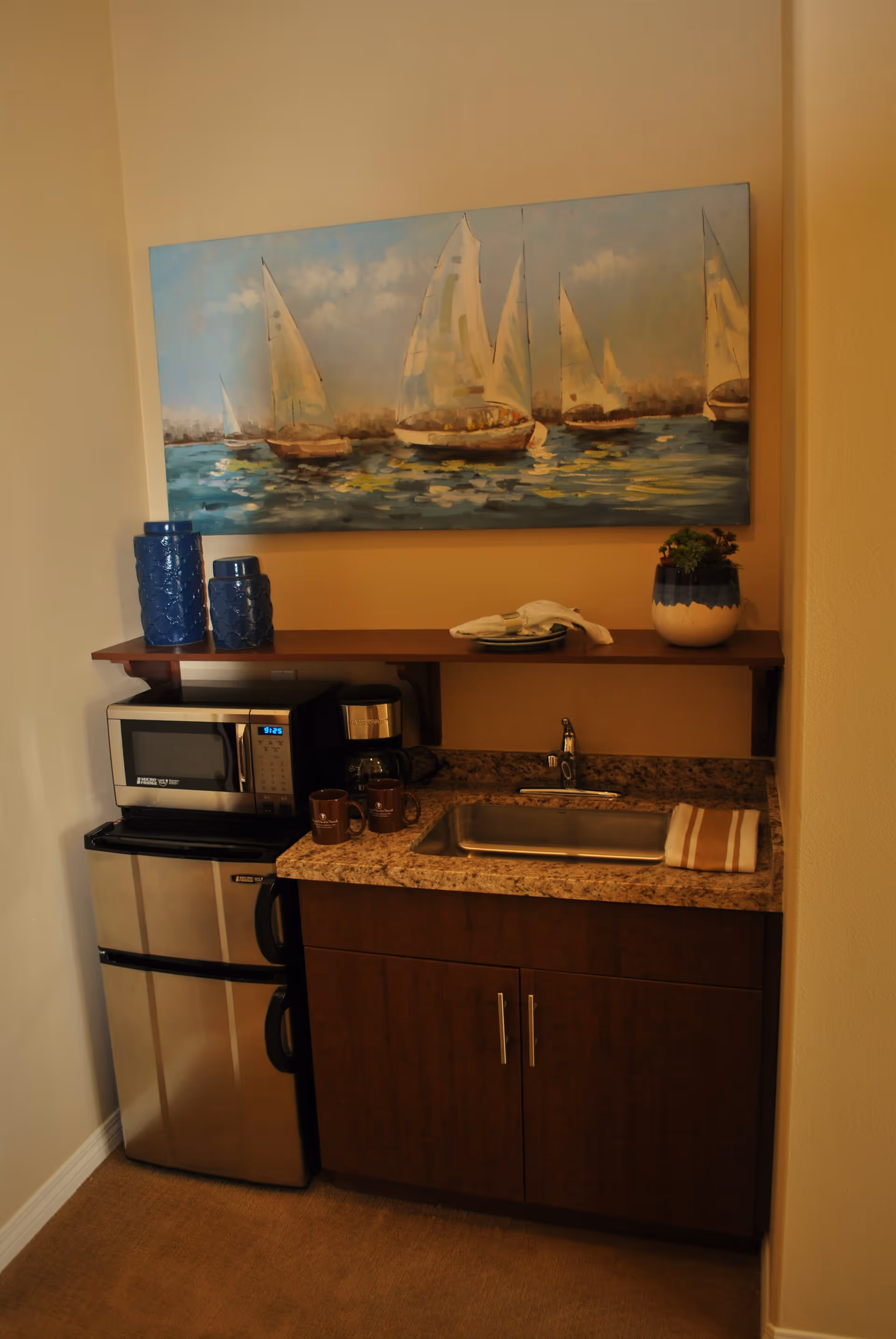A small kitchenette area with a granite countertop, stainless steel sink, and dark wooden cabinets. On the countertop are two brown mugs and a striped towel. Above the counter is a wooden shelf holding two blue decorative vases and a small potted plant. A microwave and a coffee maker sit on top of a compact stainless steel refrigerator. A large painting of sailboats on water hangs on the wall above the shelf.