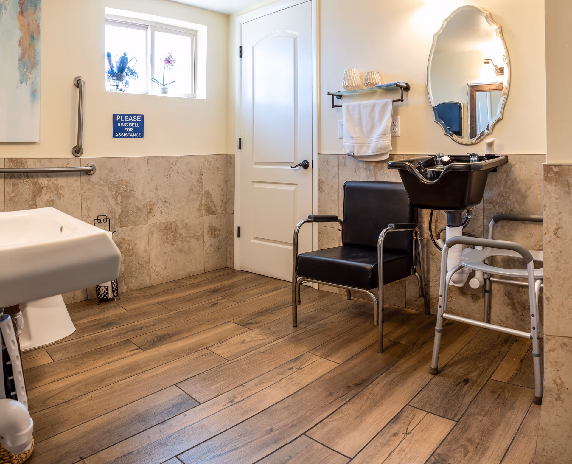 A well-lit room with a black salon chair and a black hair washing sink attached to the wall. There is a white towel hanging on a rack above the sink, a mirror with a decorative frame, a white door, and a small window with a plant on the sill. The floor is wooden, and the lower half of the walls are tiled. A walker is positioned near the sink, and a sign on the wall reads 'PLEASE RING BELL FOR ASSISTANCE.'