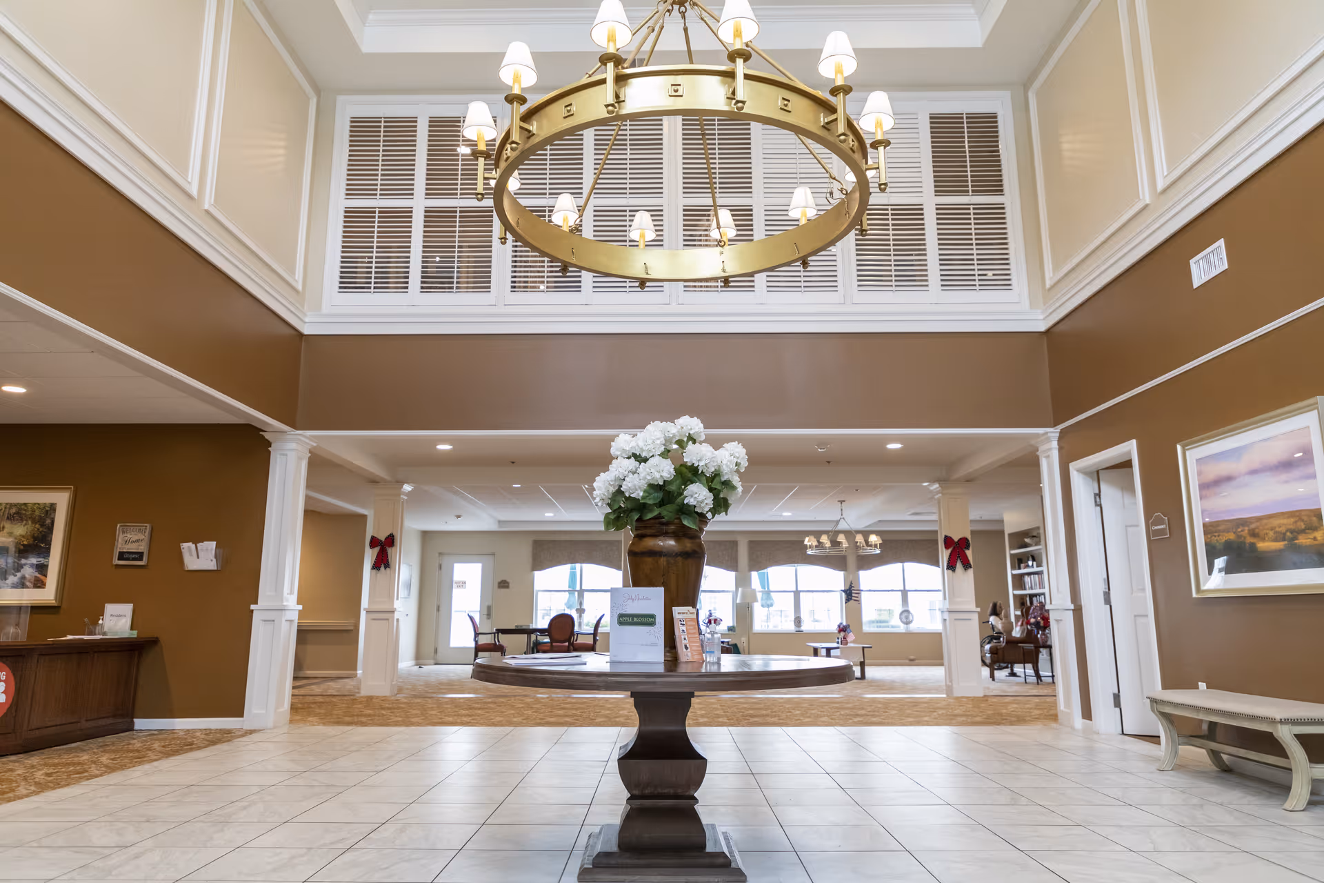 Spacious senior living facility lobby with a large round wooden table in the center holding a vase of white flowers. The room features high ceilings with a large circular chandelier, brown and white walls, tiled floor, and seating areas in the background near large windows.
