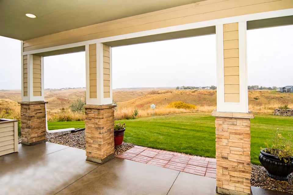 Covered patio area with stone pillars and a concrete floor overlooking a green lawn and open landscape with hills in the distance under an overcast sky.
