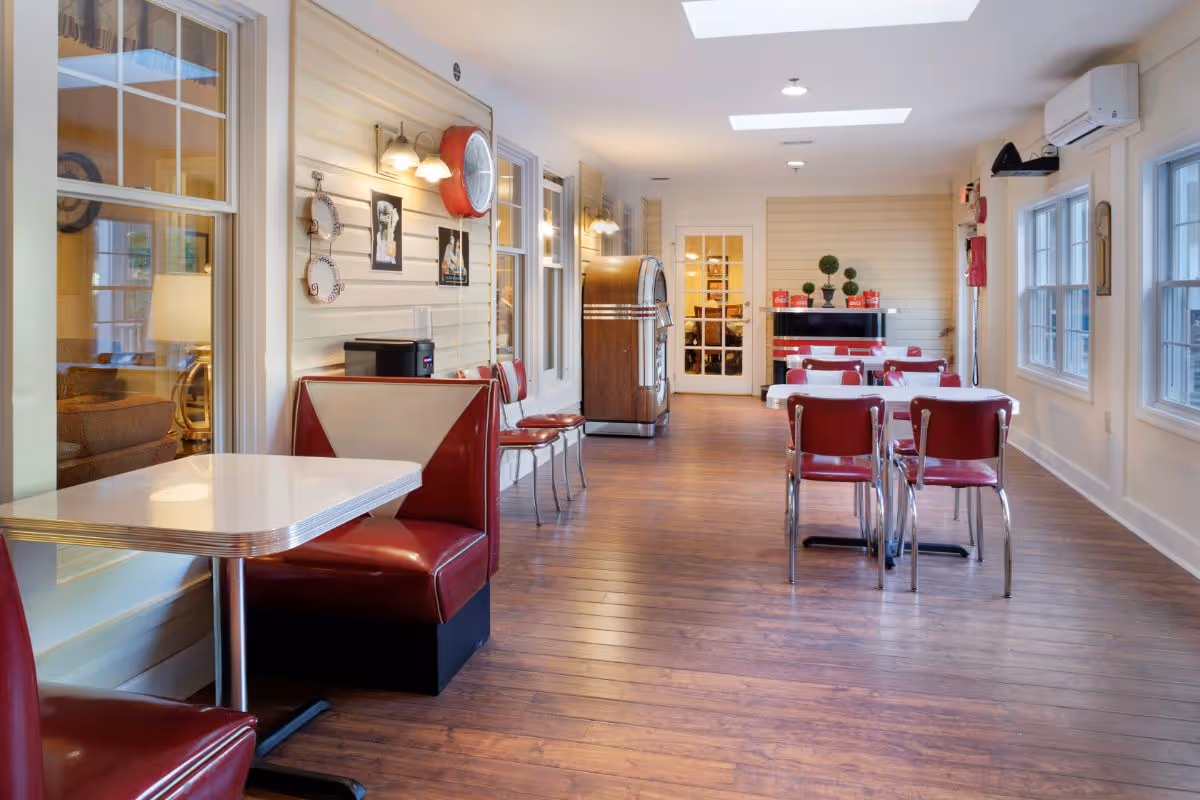 A bright dining area with retro-style red and white booths and chairs. The room has wooden flooring, cream-colored walls with decorative plates and pictures, and a vintage jukebox near the back. There are multiple windows on the right side letting in natural light, and a small counter with decorative plants and Coca-Cola cans at the far end.