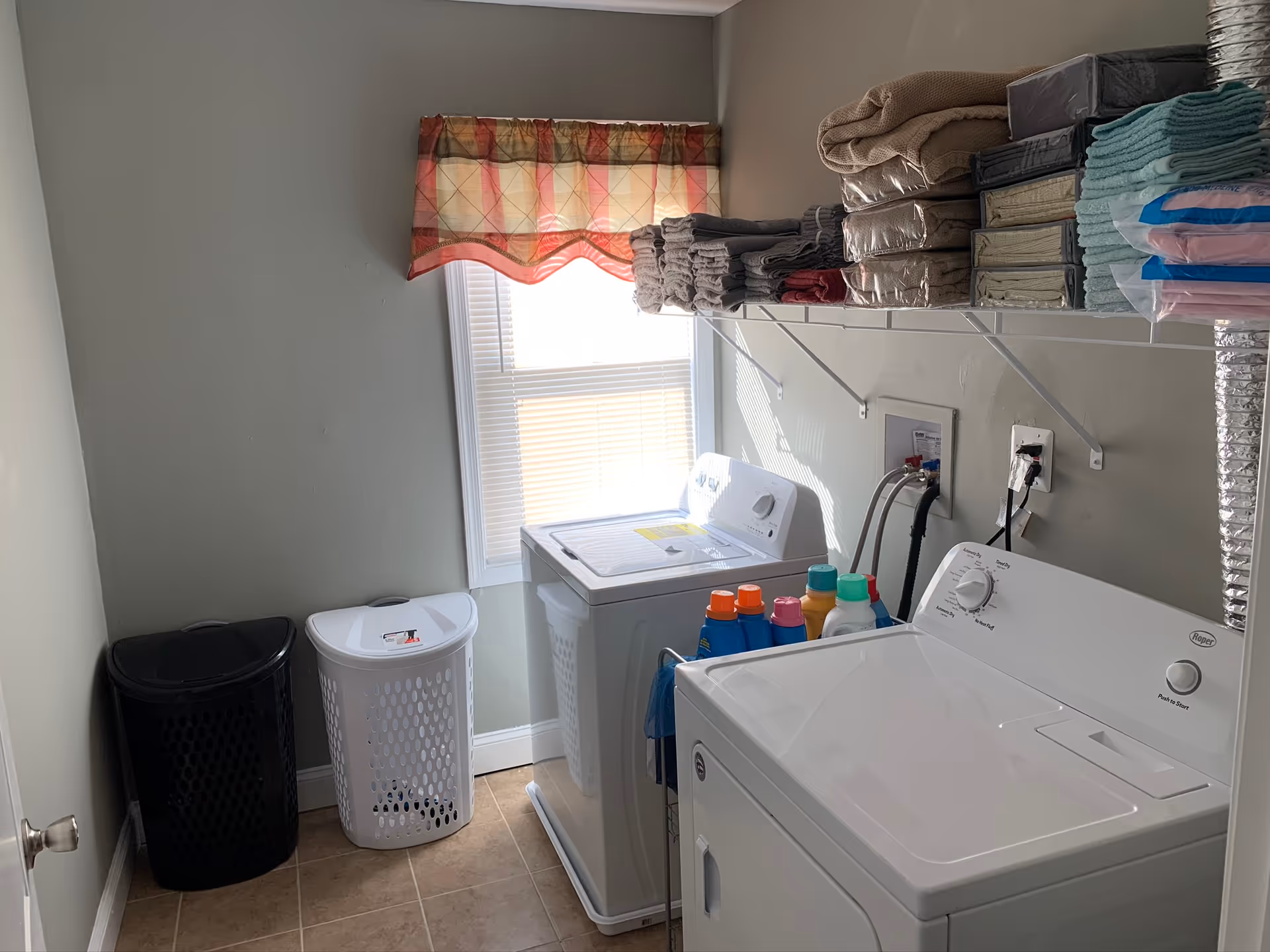 Laundry room with a washing machine and dryer side by side. Above them is a white wire shelf holding neatly folded towels and linens. Two laundry baskets, one black and one white, are placed on the floor near the wall. A window with a patterned valance allows natural light into the room.