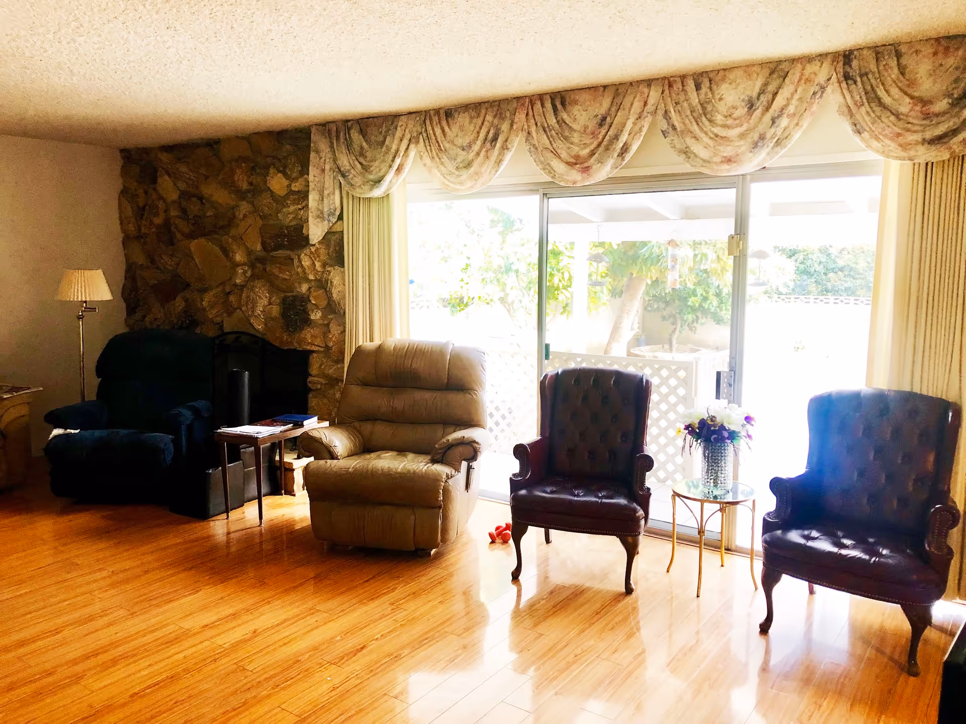 A cozy living room with a wooden floor, featuring a stone fireplace on the left side. There are four armchairs arranged in a row facing the camera: a dark blue recliner, a beige recliner, and two dark brown leather chairs with tufted backs. A small glass-top side table with a flower vase is placed between the two leather chairs. Large sliding glass doors with floral valances and curtains allow natural light to enter the room, showing a glimpse of an outdoor garden area.