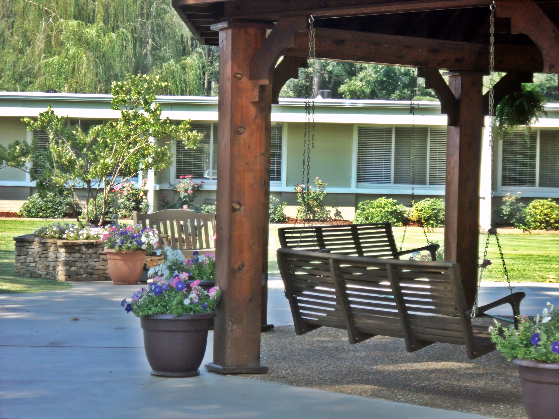 Outdoor seating area with wooden porch swings hanging from a wooden pergola. There are potted flowers and a stone planter with greenery and flowers. In the background, there is a single-story building with windows and bushes.