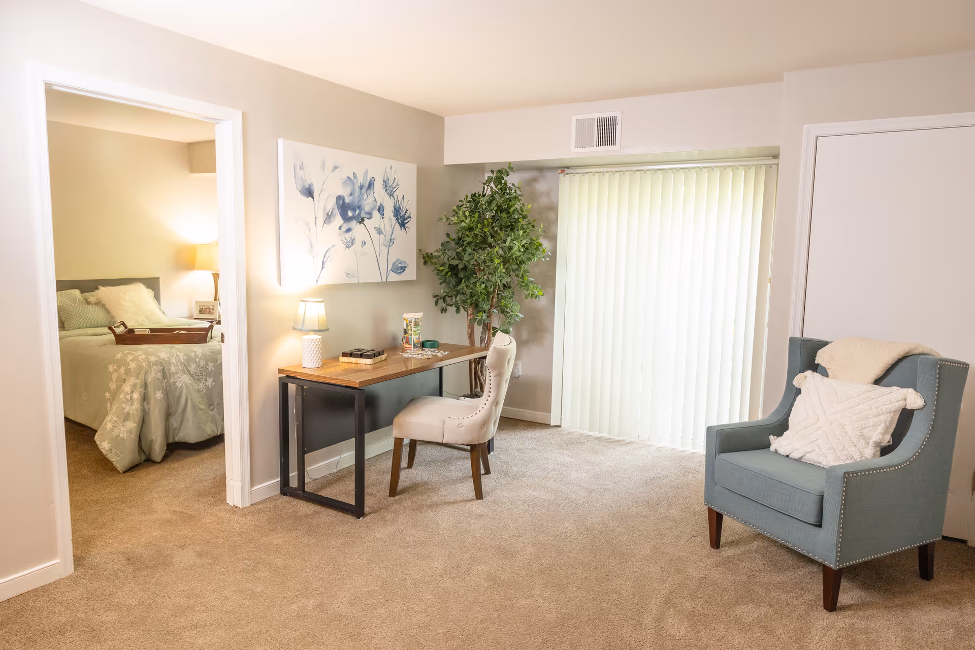 A cozy living area with beige carpet, a wooden desk with a lamp and chair, a large potted plant, and a blue armchair with a white pillow and throw. To the left, an open doorway reveals a bedroom with a bed, bedside lamp, and soft bedding. Vertical blinds cover a sliding glass door in the background.