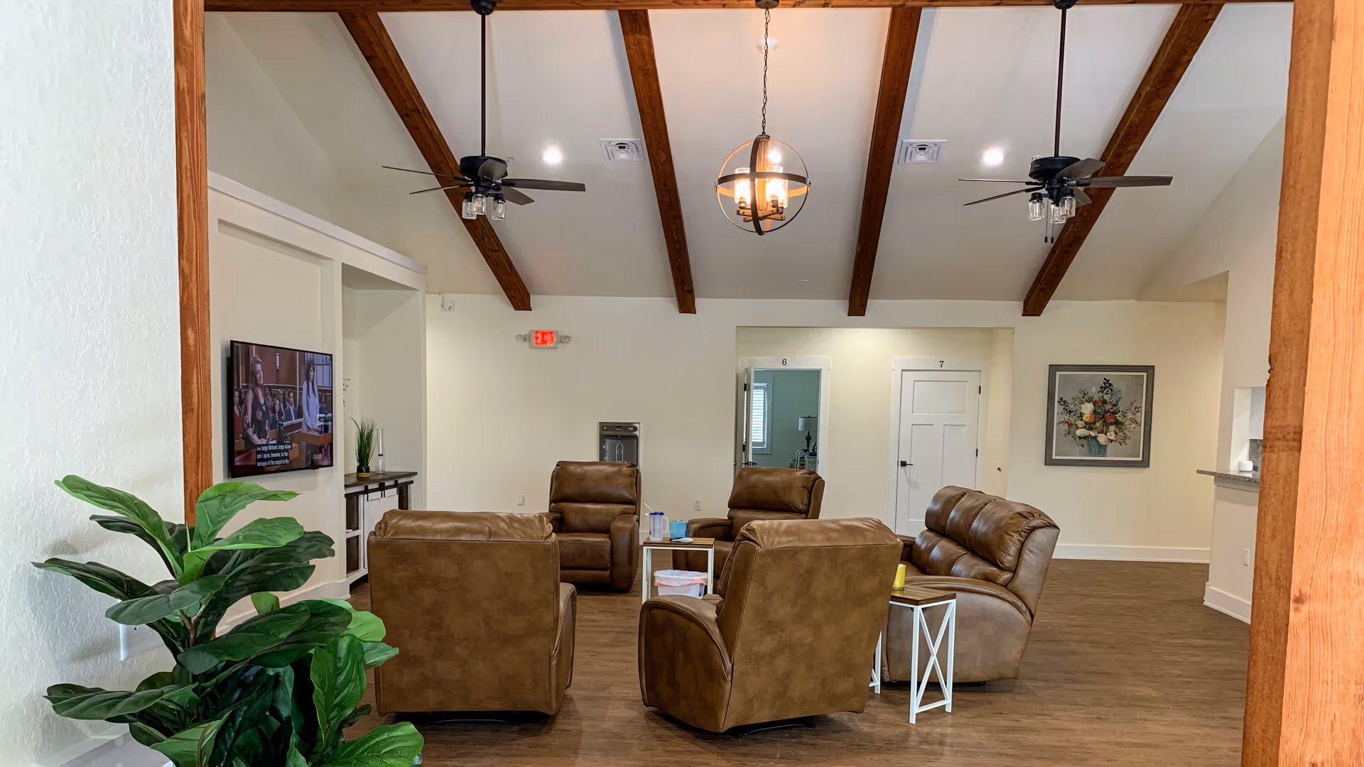 A cozy living room with four brown leather armchairs arranged around a small table. The room has wooden ceiling beams, two ceiling fans, and a hanging light fixture. A flat-screen TV is mounted on the left wall, and there is a green plant in the foreground. Doors and a framed floral picture are visible on the far wall.
