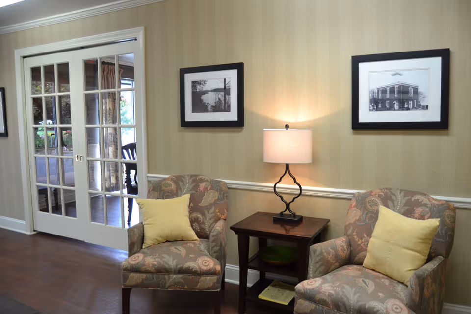 A cozy sitting area with two patterned armchairs each with a yellow pillow, separated by a wooden side table with a decorative lamp. The wall behind has two framed black and white photographs. To the left, there are glass-paneled double doors leading to another room with visible chairs and a table.