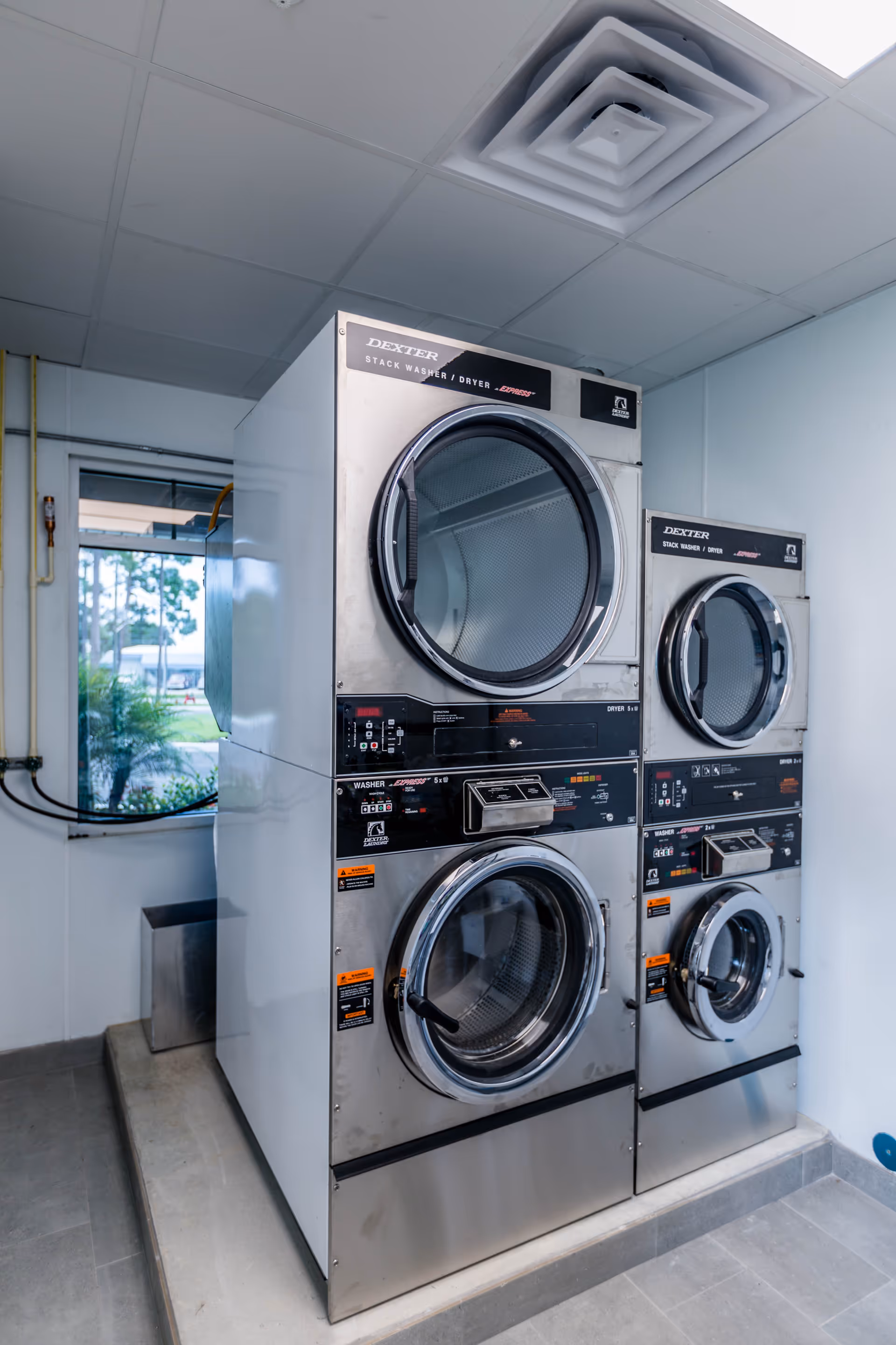 Two stacked commercial Dexter brand washer and dryer machines in a laundry room with a window showing outdoor greenery.