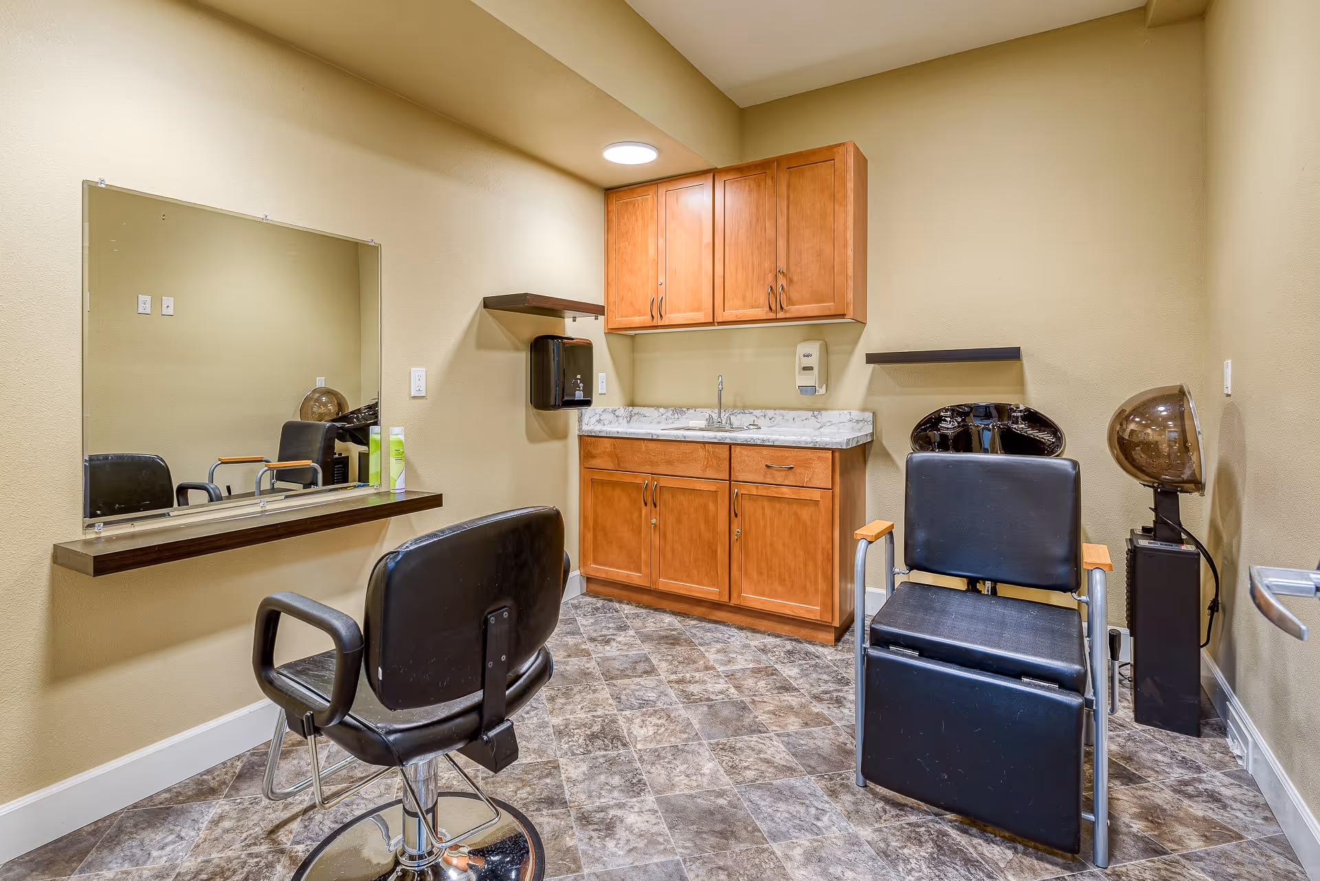 Interior view of a small salon or grooming room with two black salon chairs, a large mirror mounted on the wall, wooden cabinets with a marble countertop and sink, a hair dryer hood, and a soap dispenser mounted on the wall.