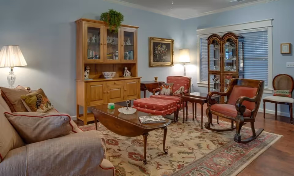 A cozy living room with a beige sofa adorned with patterned pillows, a wooden coffee table with a magazine and decorative items, a red upholstered armchair with matching ottoman, a wooden rocking chair, and a wooden cabinet with glass doors displaying decorative items. The room has a floral area rug, a floor lamp, a framed painting on the wall, and windows with blinds.