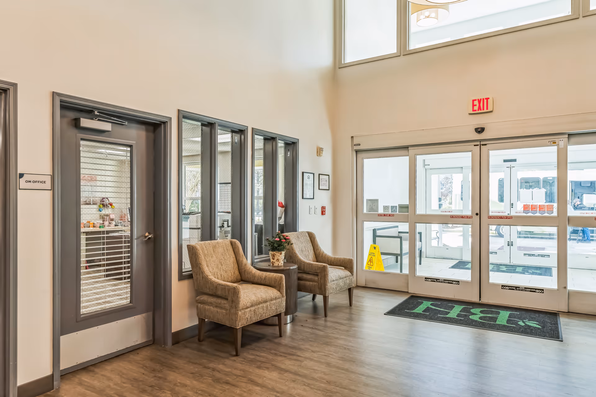 Bright senior living facility lobby with two upholstered chairs beside a small table, glass office windows and double automatic entrance doors.