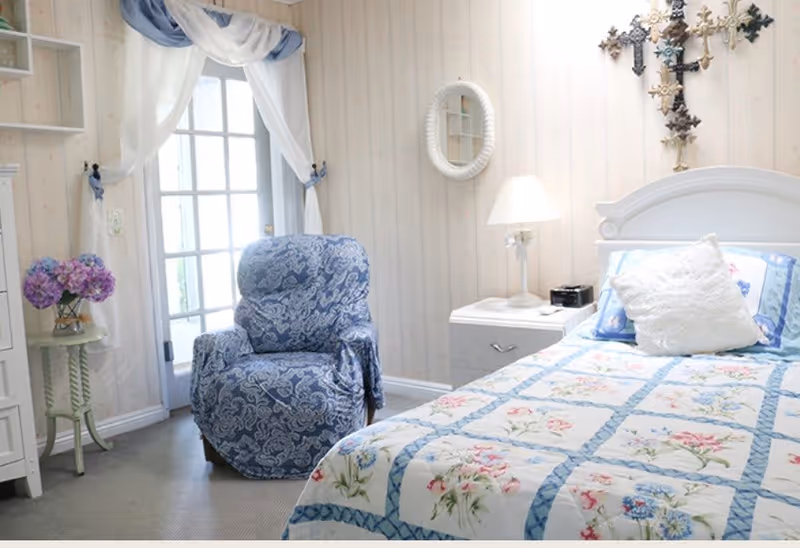 A cozy bedroom with a single bed covered in a floral quilt, a white pillow, and a blue pillow. Next to the bed is a white nightstand with a lamp and a small black clock. A blue patterned armchair is positioned near a glass door with white curtains tied back with blue fabric. A small round table with a vase of purple flowers is beside the armchair. The walls have light-colored wallpaper with subtle patterns, and decorative crosses hang above the bed.