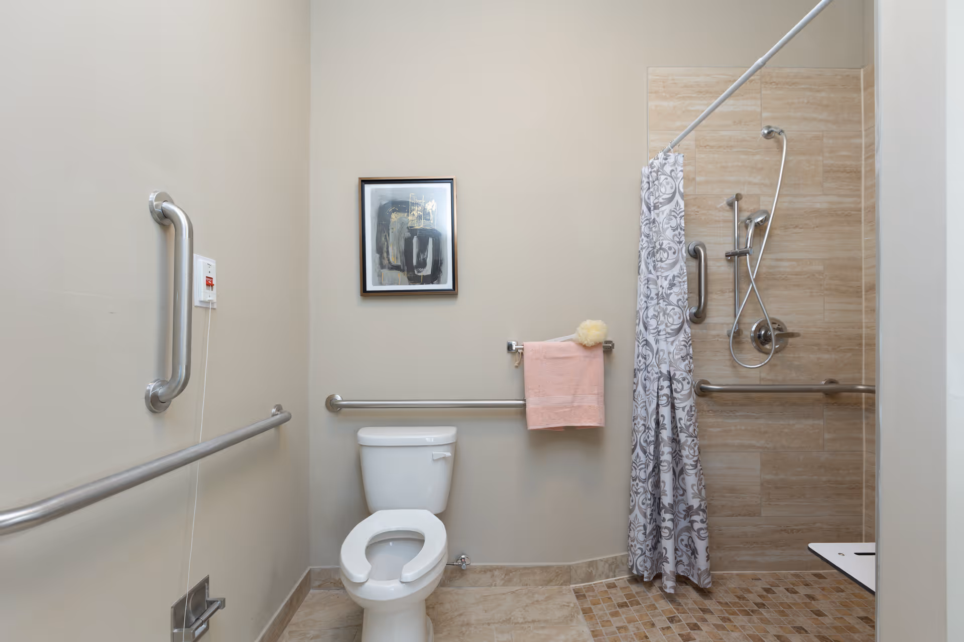 Accessible bathroom with a toilet, multiple stainless grab bars, a pink towel, framed wall art, and a tiled roll-in shower with a patterned curtain and handheld showerhead.