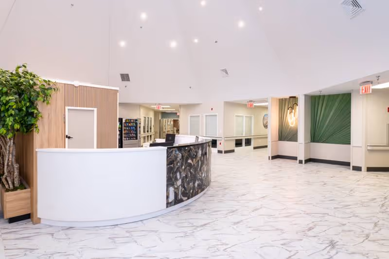 A spacious and modern reception area with a curved white and dark marble front desk, white marble-patterned flooring, and a high ceiling with recessed lighting. There is a large potted plant to the left of the desk, green decorative wall panels on the right, and multiple doorways and hallways leading to other parts of the facility.