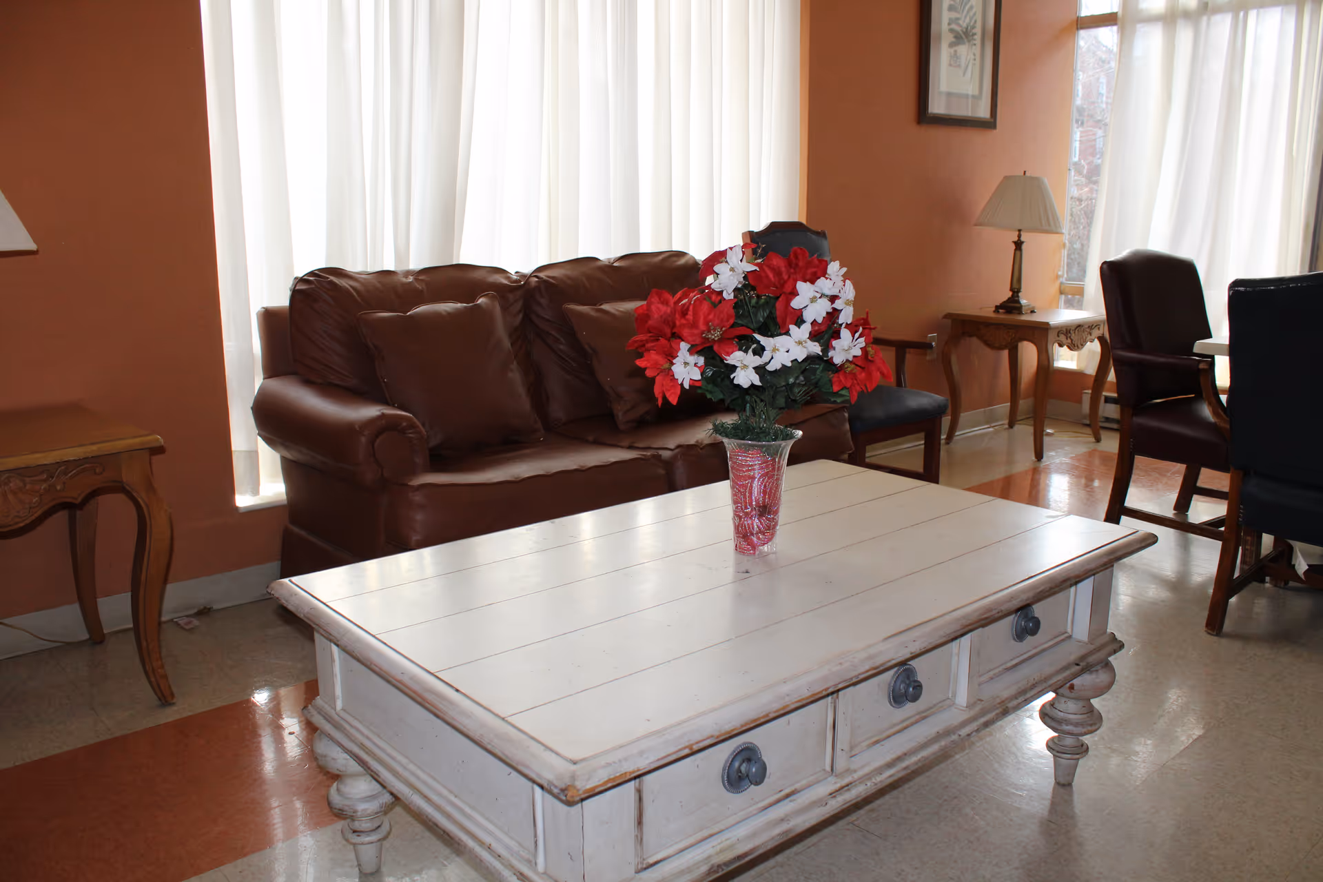 A cozy living room area with a brown leather sofa, a white wooden coffee table with drawers, and a vase of red and white flowers on the table. There are two side tables with lamps, a framed picture on the wall, and several chairs near a window with vertical blinds.