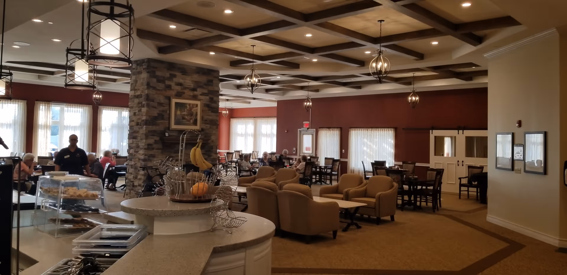 A spacious dining and lounge area in a senior living facility with multiple tables and chairs, comfortable armchairs, a stone pillar with a framed picture, pendant lights hanging from a coffered ceiling, and large windows with sheer curtains allowing natural light to fill the room. Several elderly residents and a staff member are visible in the background.