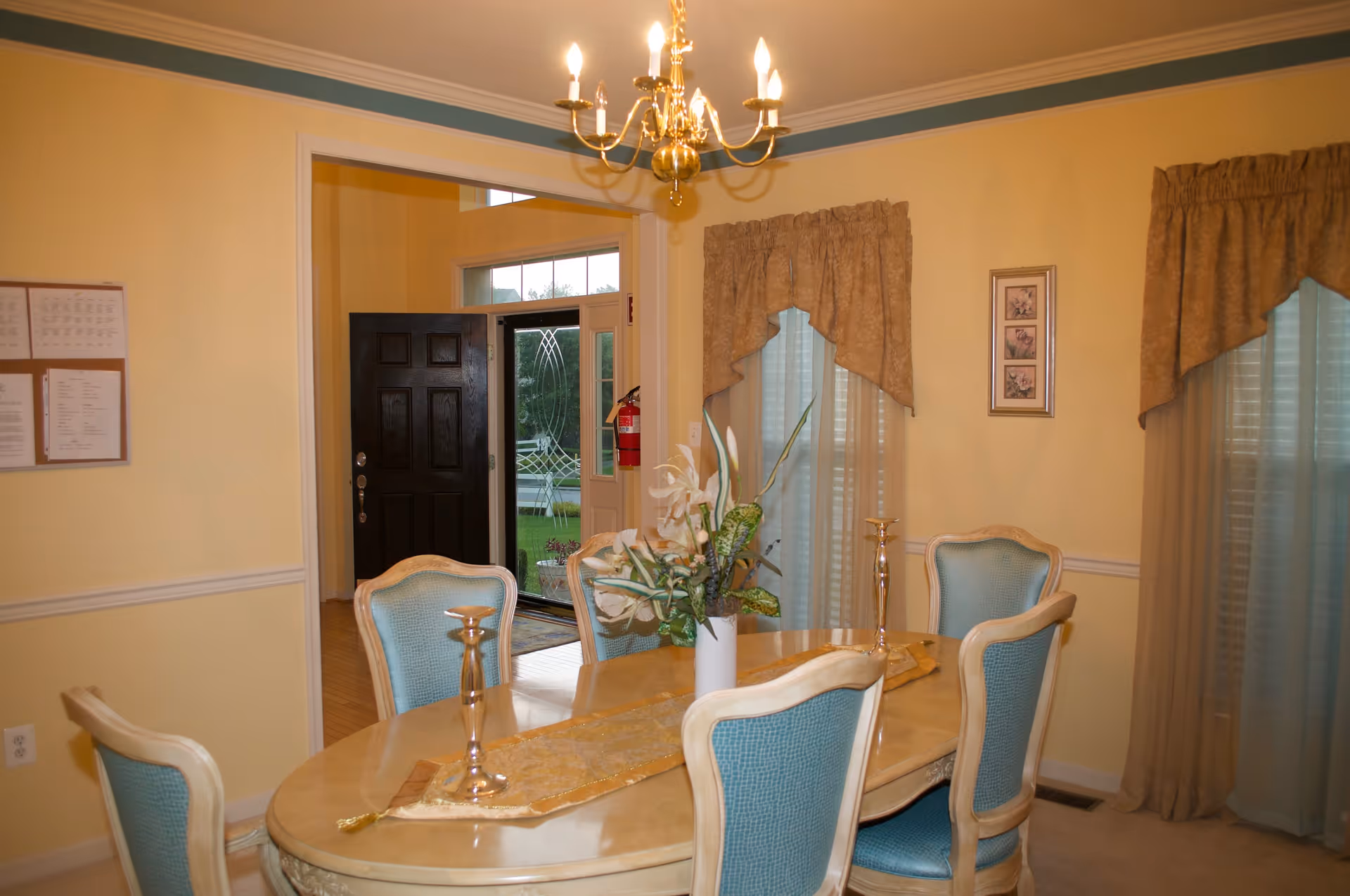 A dining room with a light wood oval table surrounded by six chairs with blue upholstery. The table has a gold runner, two silver candlesticks, and a white vase with artificial flowers. The room has yellow walls with white trim, two windows with beige curtains, and a gold chandelier hanging from the ceiling. In the background, there is an open black front door with glass panels and a fire extinguisher mounted on the wall.