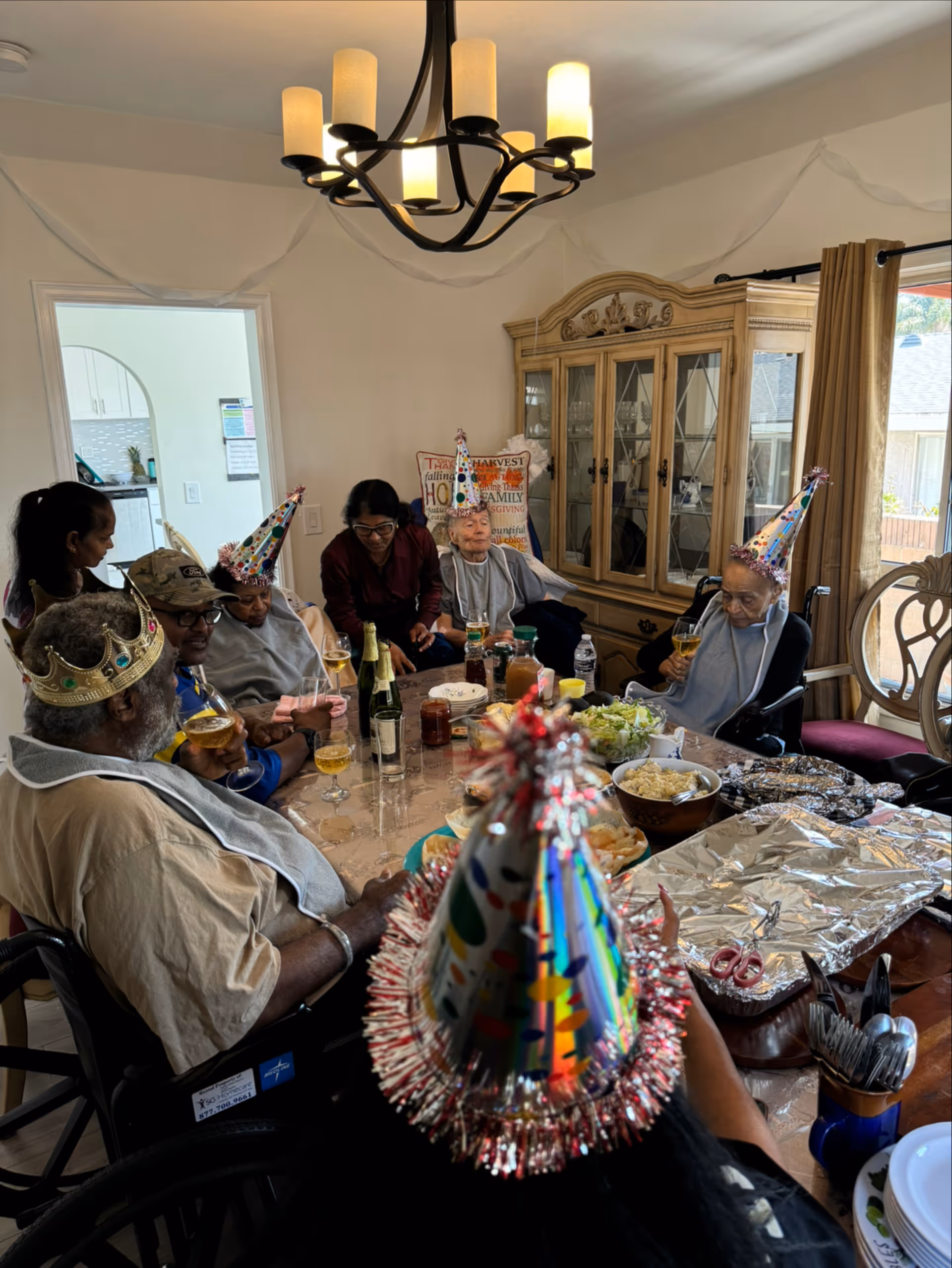 A group of seniors in party hats celebrating around a dining table with food and drinks.