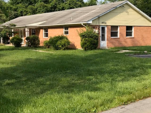 Single-story brick building with a light-colored roof and a small front porch, surrounded by green grass and some bushes. The building has several windows and a door with the number 1571 above it.