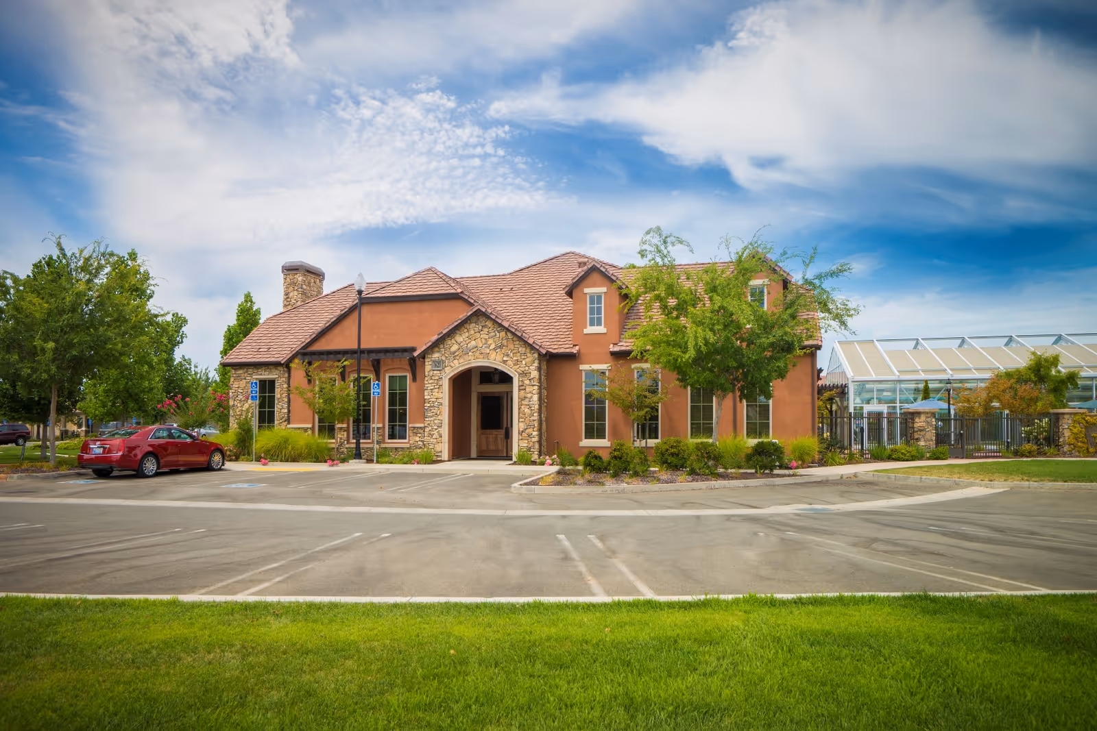 Front exterior view of a single-story building with a stone and stucco facade, surrounded by trees and landscaping. There is a parking lot with a red car parked in a handicapped spot. The sky is partly cloudy with blue patches.
