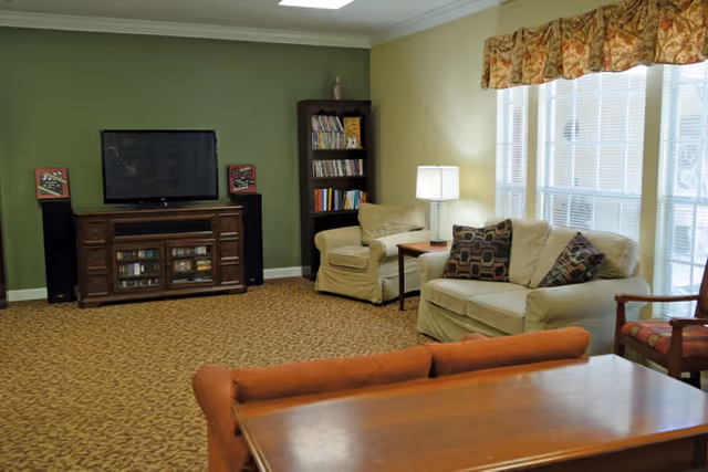 A cozy living room with a green accent wall featuring a flat-screen TV on a wooden cabinet flanked by two speakers. There is a beige armchair and sofa with patterned pillows near a window with floral valances. A wooden bookshelf filled with books is in the corner, and a wooden table with an orange cushioned bench is in the foreground.