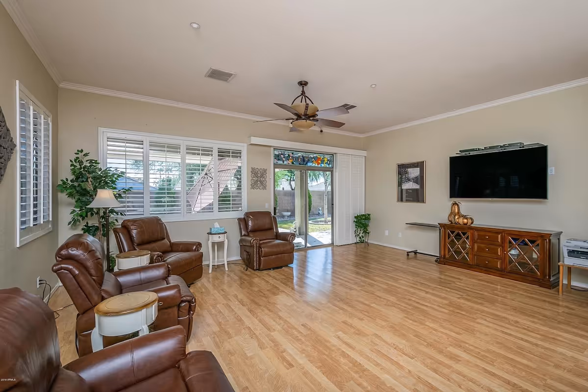 A spacious living room with light wood flooring, beige walls, and a ceiling fan. The room features four brown leather recliners arranged along the left side and back wall, with small round side tables between them. Large windows with white shutters and a sliding glass door provide natural light and a view of the outdoor area. A wooden media console with glass-paneled doors sits against the right wall beneath a mounted flat-screen TV. There are a couple of green plants and framed artwork on the walls.
