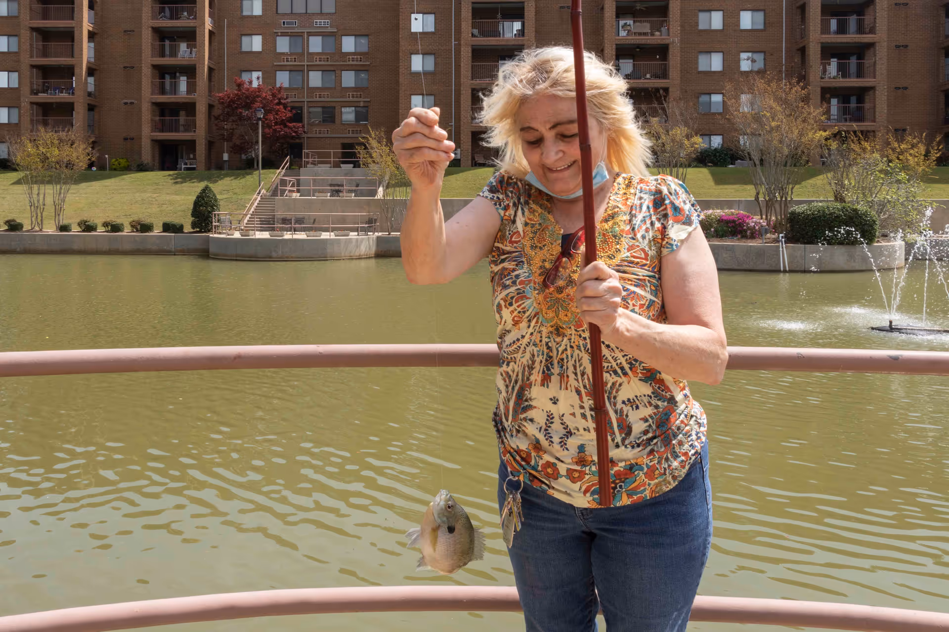 A woman standing on a railing by a pond holding a fishing rod with a fish caught on the line. Behind her is a large multi-story brick building with balconies and landscaped greenery around the pond.