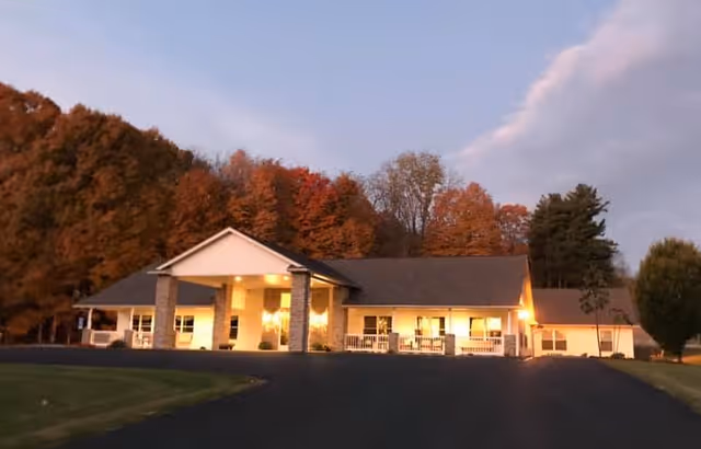 Single-story assisted living building with a lit covered entrance, driveway, and autumn trees in the background at dusk.