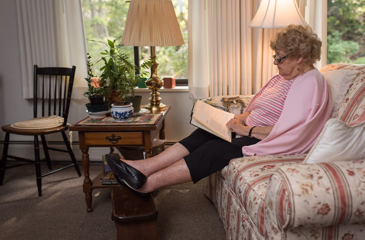 An elderly woman wearing glasses and a pink shawl is sitting on a floral patterned couch reading a book. Her feet are resting on a small wooden footstool. Next to her is a wooden side table with several potted plants and a brass lamp. A wooden chair with a woven seat is positioned nearby, and a window with white curtains lets in natural light.