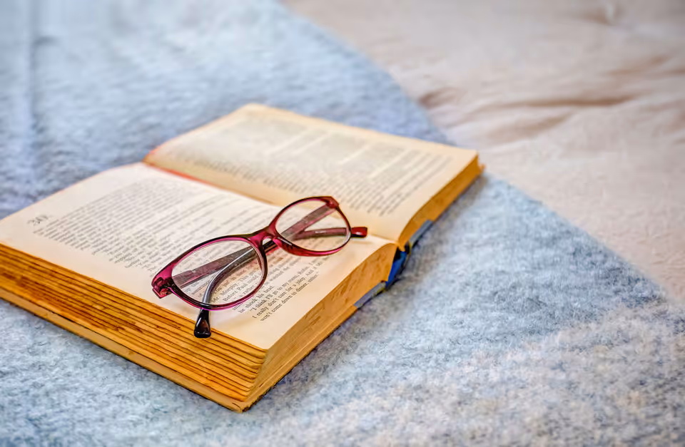 An open book with yellowed pages resting on a soft, light blue blanket, with a pair of red-framed eyeglasses placed on top of the book.